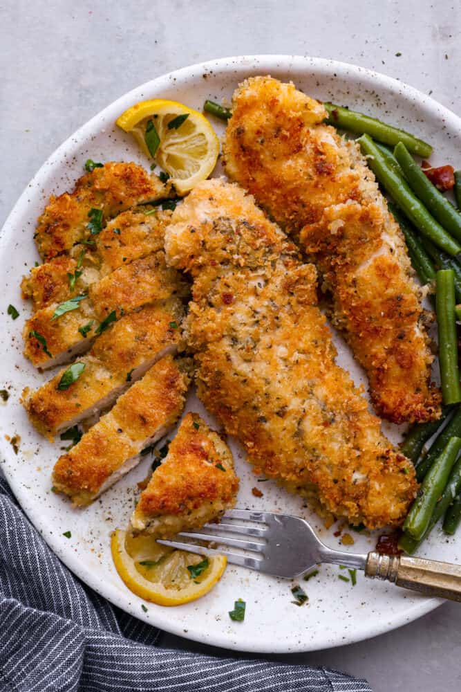 Top view of chicken Milanese on a white plate. One of the cutlets is sliced with a fork on the plate. Green beans and lemon slices are next to the chicken.