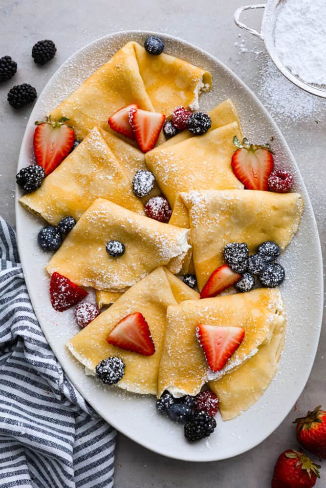 Overhead view of folded crepes on a gray platter dusted with powdered sugar and topped with fresh berries.