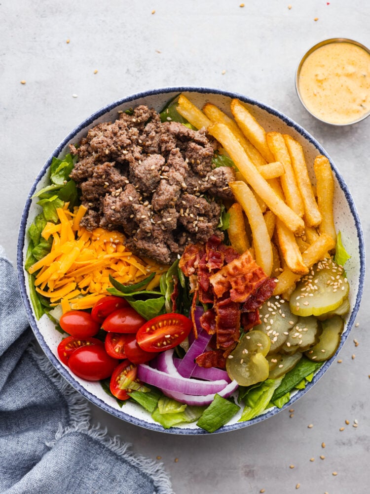 Top-down view of a blue and white bowl filled with ground beef, onions, pickles, tomatoes, lettuce, French fries, and bacon, topped with Big Mac sauce.