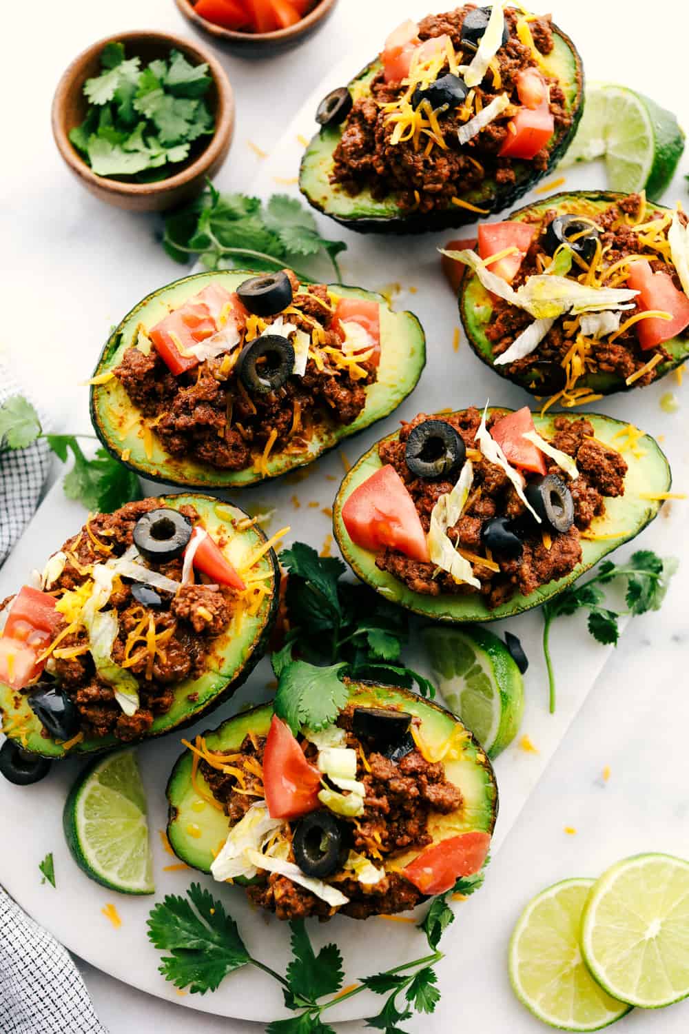 Top view of taco stuffed avocados on a white marble slab. The avocados are garnished with tomatoes, olives, cheese, and cilantro.