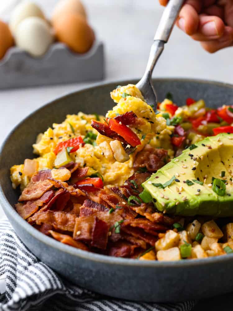 Close view of a fork lifting up eggs, bacon, and veggies from the breakfast bowl.