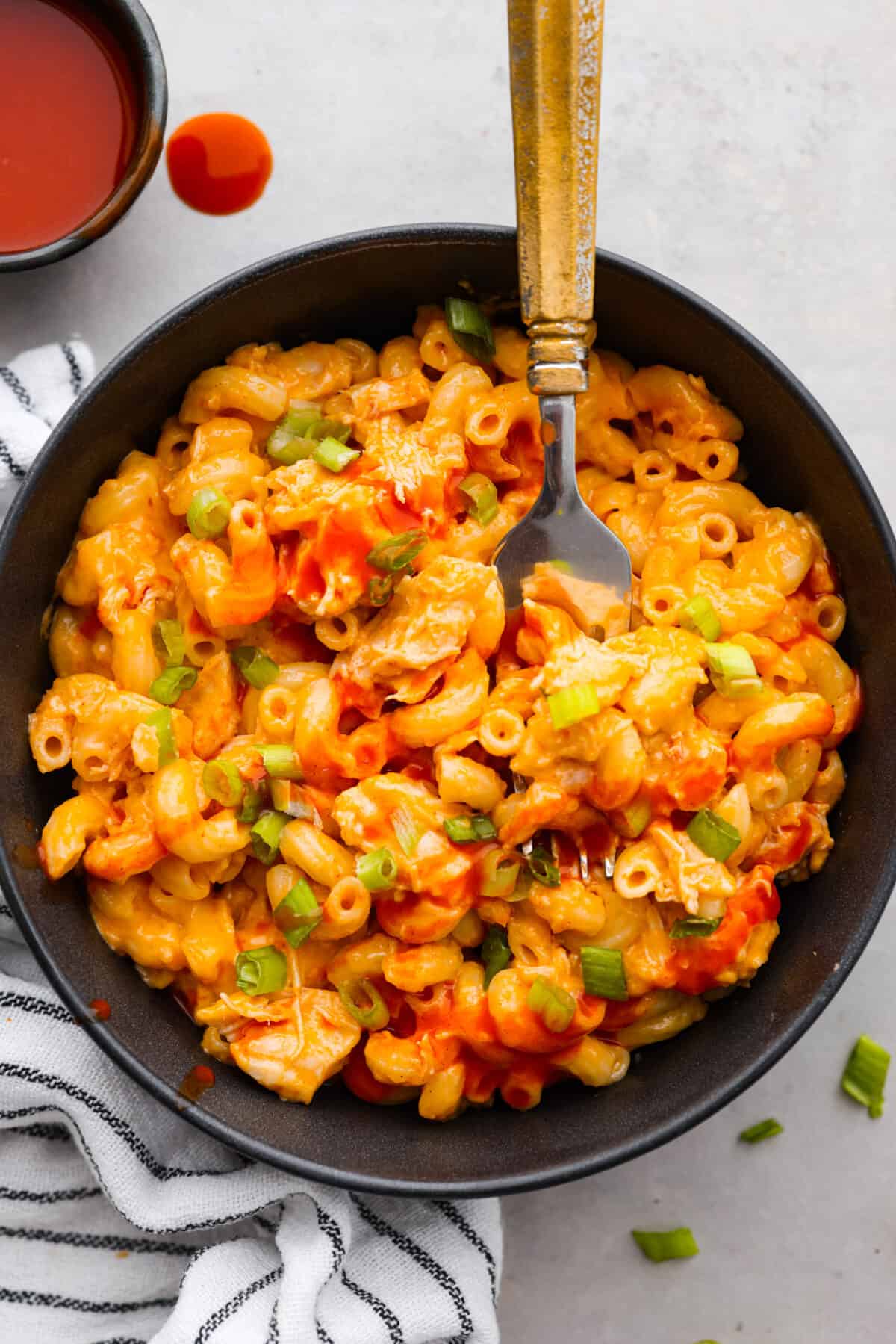 Top view of buffalo chicken mac and cheese in a black bowl with a silver fork. Hot sauce is drizzled on top and in a bowl on the side.