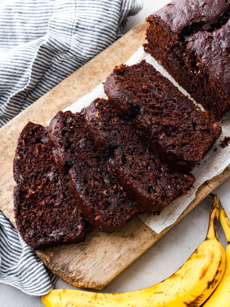 Top view of sliced chocolate banana bread on a wood cutting board. Bananas and a striped towel are next to the cutting board.