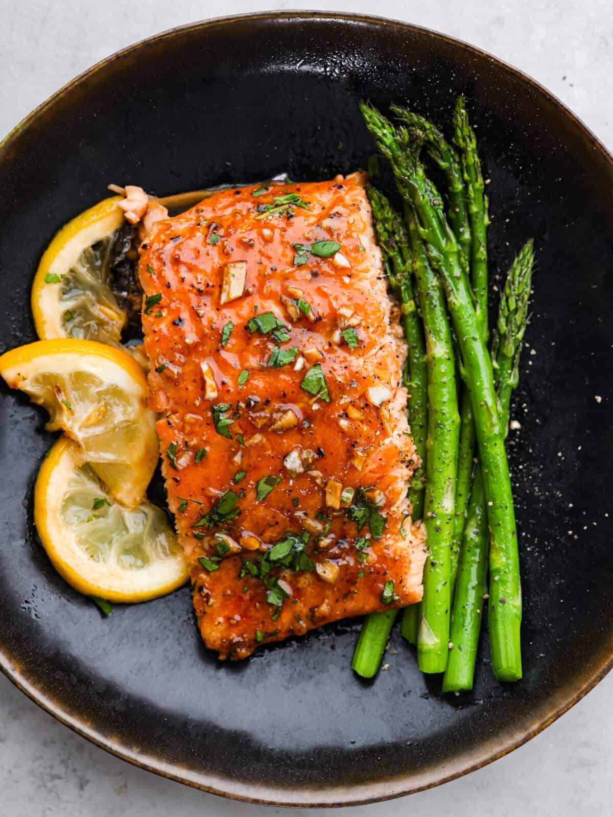 A black plate with garlic brown sugar salmon, lemon slices and asparagus. 