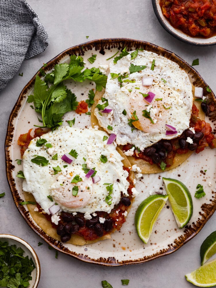 Top view of huevos rancheros on a tan and brown plate. Garnished with cilantro, limes, red onions and cheese.