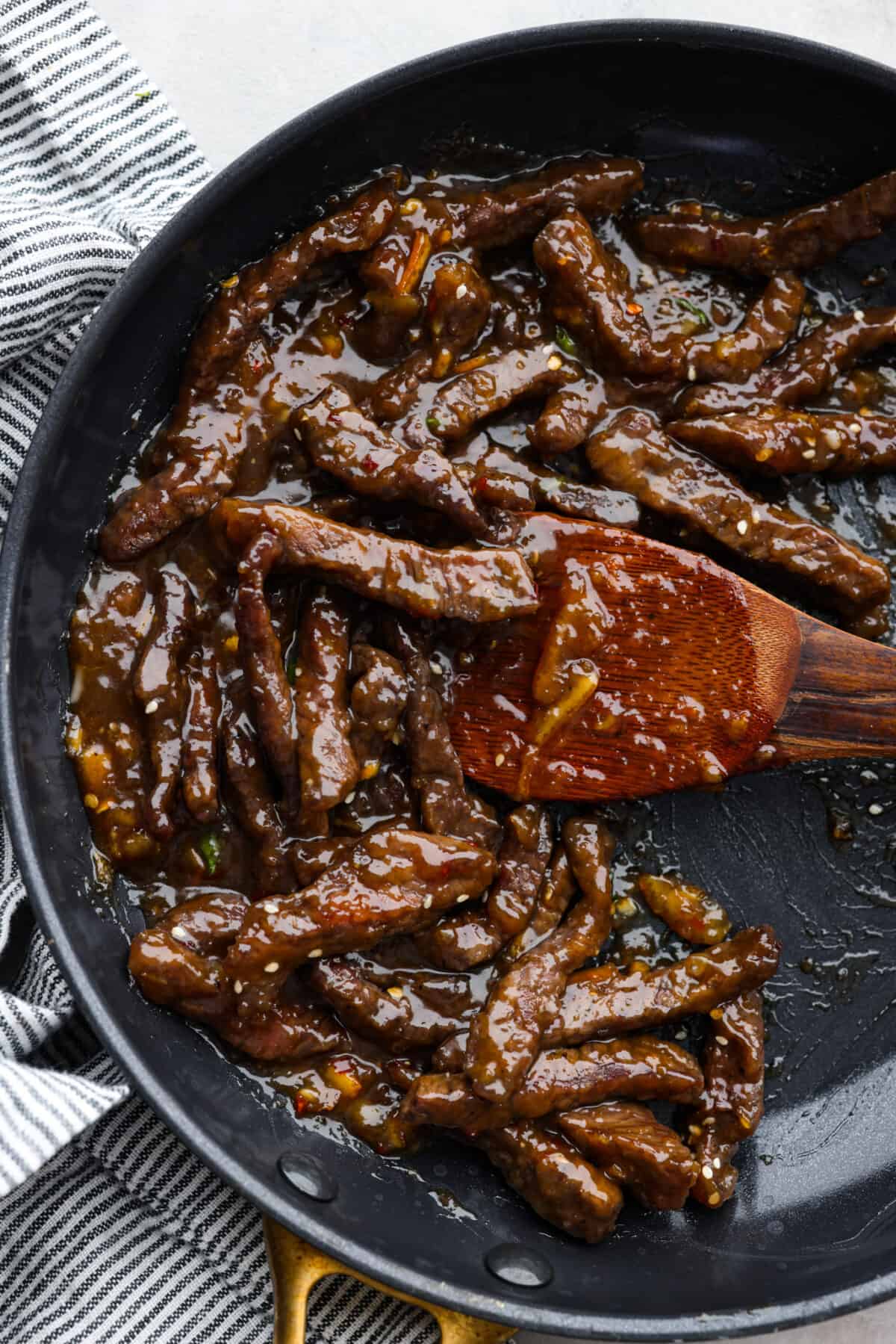 Top-down view of orange beef in a skillet, being stirred with a wooden spoon.