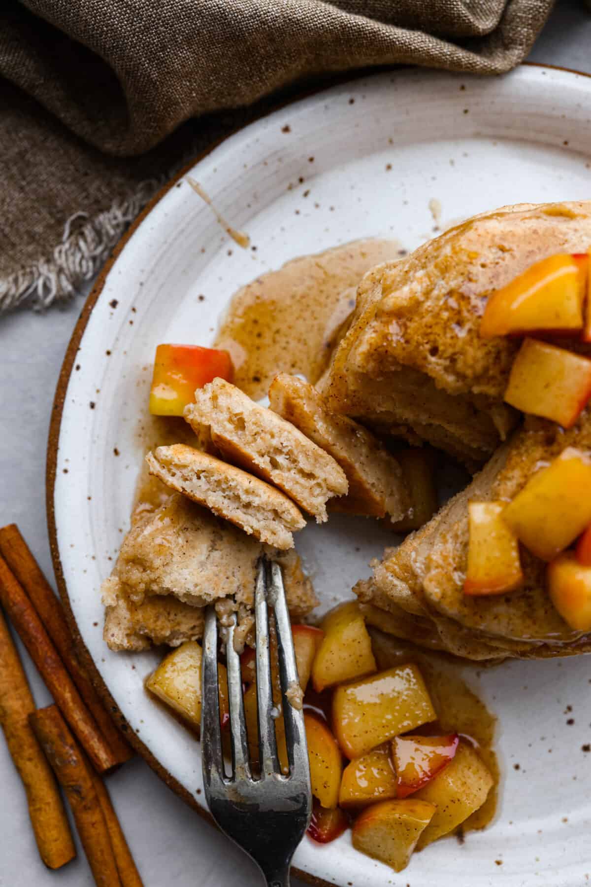 Top view of a fork cutting into applesauce pancakes. Sauteed apples and syrup are on the plate of pancakes.