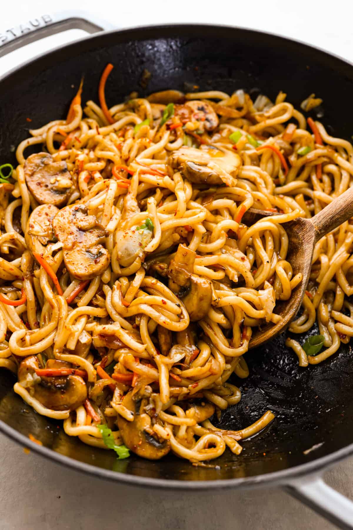 Top close view of udon noodles in a large black wok and a wooden spoon.
