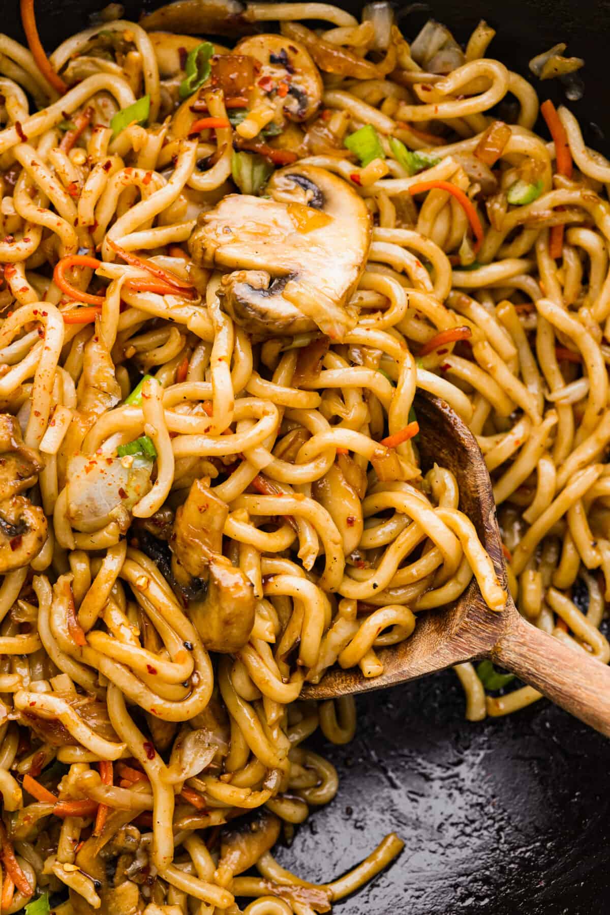Close view of noodles in a wok with a wood spoon lifting up noodles.