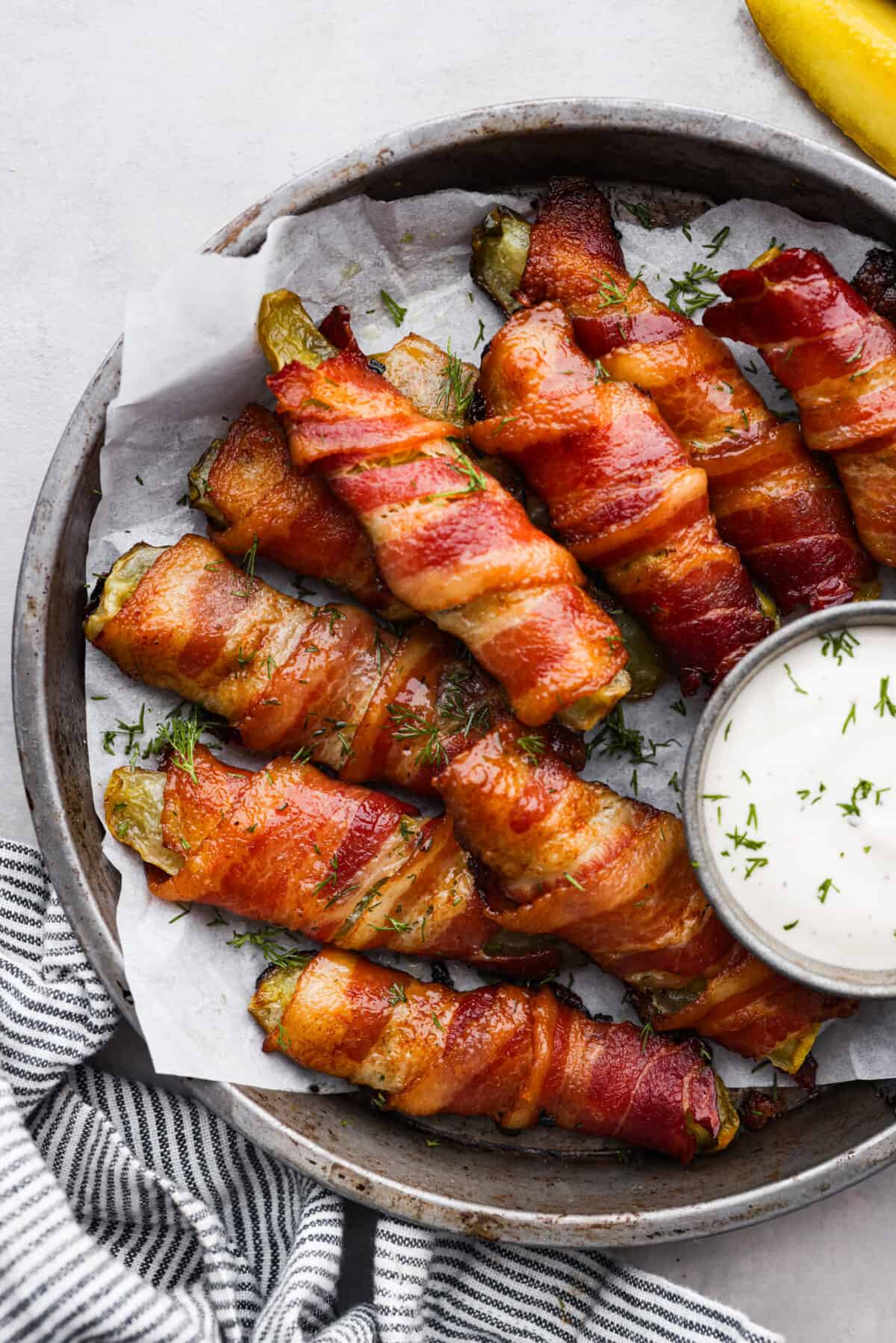 Bacon wrapped pickles in a gray serving bowl, served with a side of ranch dressing.