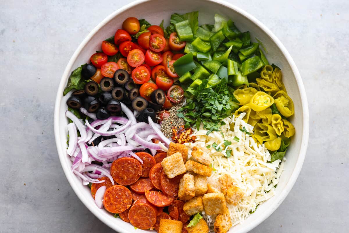 The top view of a bowl with lettuce and ingredients that aren't mixed yet. Cheese, croutons, pepperonis, peppers, olives, tomatoes, onions and herbs and spices. 