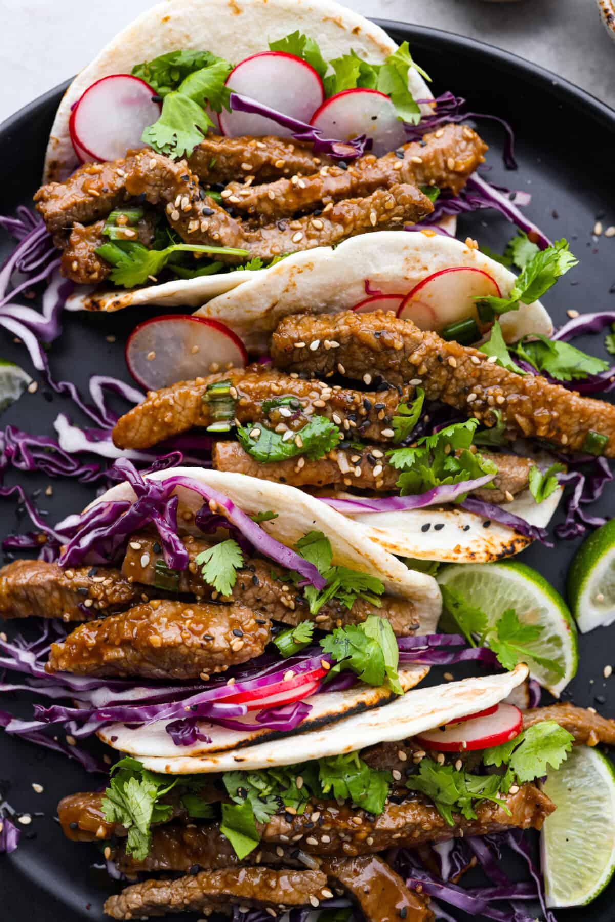 Close view of tacos on a black plate garnished with cabbage, cilantro, radishes, limes, and sesame seeds.