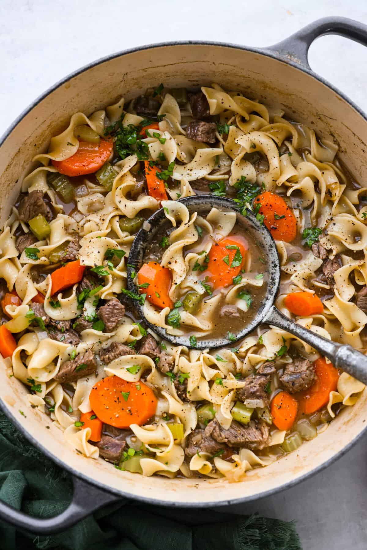 Beef noodle soup being scooped up with a ladle in a large pot.