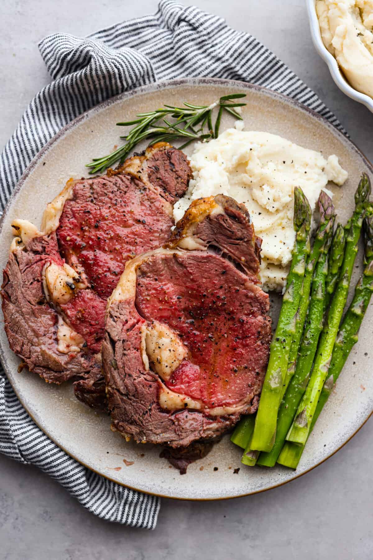 Top view of sliced ribeye roast on a large plate with mashed potatoes and asparagus on the side.