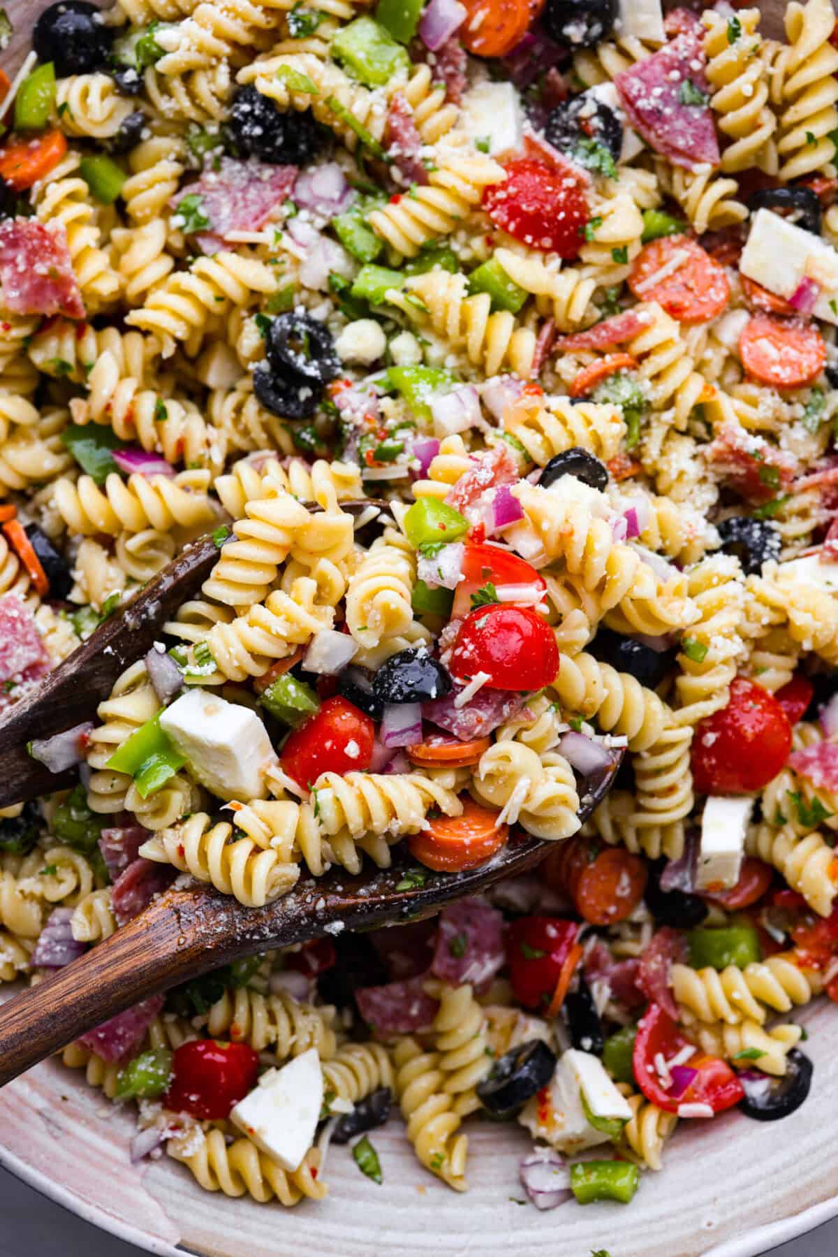 Close up shot of pasta salad with rotini noodles, tomatoes, bits of red onion, olives, salami, green pepper and cheese. 